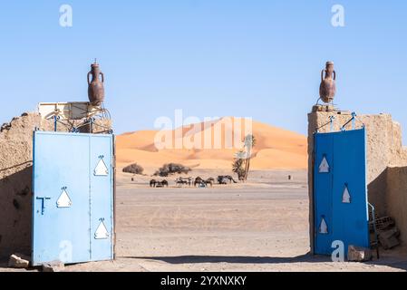 Blick auf die Dünen der Merzouga-Wüste in Marokko von der Eingangstür zu einem Riad. Eine kleine Palmenplantage ist auf dem trockenen Land zu sehen und so Stockfoto