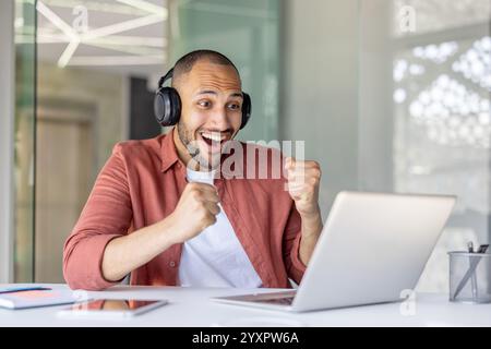 Ein fröhlicher Mann, der Kopfhörer trägt, feiert freudig eine Leistung, während er an seinem Laptop arbeitet. Begeisterung und positive Energie in einer modernen Büroumgebung erfassen. Stockfoto