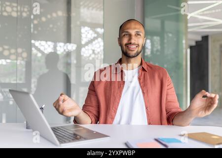 Ein junger Mann in einem lässigen Hemd, der an einem Schreibtisch mit Laptop in einer hellen und modernen Büroumgebung lächelt und meditiert. Die Stimmung vermittelt Ruhe und Gleichgewicht während der Arbeitszeit. Stockfoto