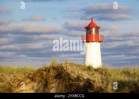 "Malerischer rot-weißer Leuchtturm, der hoch vor einem wolkengefüllten blauen Himmel steht, umgeben von windgepeitschtem Gras und Küstendünen." Stockfoto