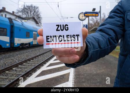 Bayern, Deutschland - 17. Dezember 2024: Eine Person hält ein Schild mit dem Reading Train entgleist vor einem Bahnhof und Gleisen. Symbolisches Bild für Zugunfälle, Störungen des Bahnverkehrs und Risiken im Schienenverkehr. FOTOMONTAGE *** eine Person hält ein Schild mit der Aufschrift öZug entgleist vor einem Bahnhof und Gleisen. Symbolbild für Zugunfälle, Bahnbetriebsstörungen und Risiken im Schienenverkehr. FOTOMONTAGE Stockfoto