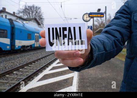 Bayern, Deutschland - 17. Dezember 2024: Eine Person hält ein Schild mit der Aufschrift Unfall vor einem Bahnhof und Gleisen. Symbolisches Bild für Zugunfälle, Bahnverkehrsstörungen und Gefahren im Schienenverkehr. FOTOMONTAGE *** eine Person hält ein Schild mit der Aufschrift öUnfall vor einem Bahnhof und Gleisen. Symbolbild für Zugunglücke, Bahnbetriebsstörungen und Gefahren im Schienenverkehr. FOTOMONTAGE Stockfoto