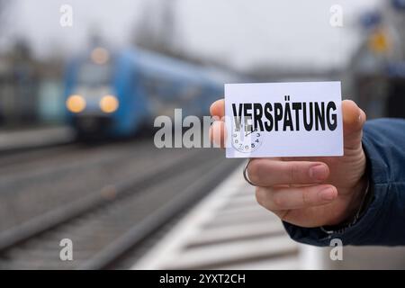 Bayern, Deutschland - 17. Dezember 2024: Eine Person hält ein Schild mit dem Text VERSPÄTUNG vor einem Bahnhof und Gleisen. Symbolisches Bild für Zugverspätungen, Verkehrsprobleme und Reisestörungen im Schienenverkehr. FOTOMONTAGE *** eine Person hält ein Schild mit dem Text VERSPÄTUNG vor einem Bahnhof und Gleisen. Symbolbild für Zugverspätungen, Verkehrsprobleme und Reiseunterbrechungen im Schienenverkehr. FOTOMONTAGE Stockfoto