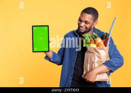 Ein selbstbewusster afroamerikanischer Mann präsentiert Grüncreen auf einer Tablette und trägt eine Tasche mit Gemüse auf gelbem Hintergrund. Zufriedenes männliches Modell mit isoliertem Modell-Display auf dem Gerät. Stockfoto