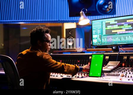 Tontechniker mischt Titel neben dem Greenscreen-Tablet im Kontrollraum, Bedienpult und Fader. afroamerikanischer Produzent, der Hits aufnimmt und Sounds verarbeitet. Stockfoto