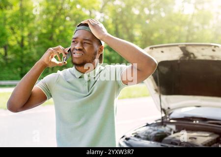 Unglücklicher schwarzer Mann, der in der Nähe seines kaputten Autos mit offener Haube steht und Pannendienst auf dem Smartphone anruft Stockfoto