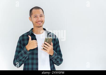 Junge asiatische Männer, die Flanell und Hemd tragen, halten das Smartphone mit den Daumen nach oben auf isoliertem weißen Hintergrund Stockfoto