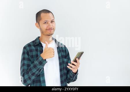 Junge asiatische Männer, die Flanell und Hemd tragen, halten das Smartphone mit den Daumen nach oben auf isoliertem weißen Hintergrund Stockfoto