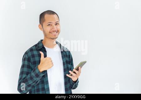 Junge asiatische Männer, die Flanell und Hemd tragen, halten das Smartphone mit den Daumen nach oben auf isoliertem weißen Hintergrund Stockfoto