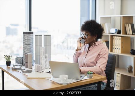 Selbstbewusste afrikanische Architektin im Büro, die auf dem Smartphone über das Stadtprojekt spricht. Profi mittleren Alters, umgeben von Wolkenkratzermodellen, arbeiten an Laptops und schaffen zukünftige Stadtentwicklung. Stockfoto