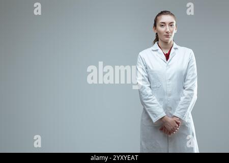 Eine junge, selbstbewusste Technikerin im Labormantel posiert in einem Studio mit einem ernsthaften Gesichtsausdruck und blickt mit ihrem han direkt in die Kamera Stockfoto