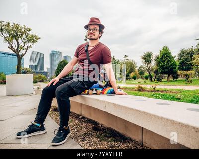 Stilvoller junger Mann mit Hut und Brille sitzt auf einer Bank mit Skateboard, im urbanen Park mit Grün und Wolkenkratzern. Stockfoto