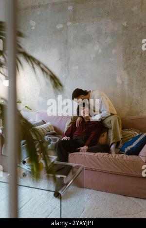 Liebevoller schwuler Mann, der einen Freund küsst, der zu Hause auf dem Sofa sitzt Stockfoto