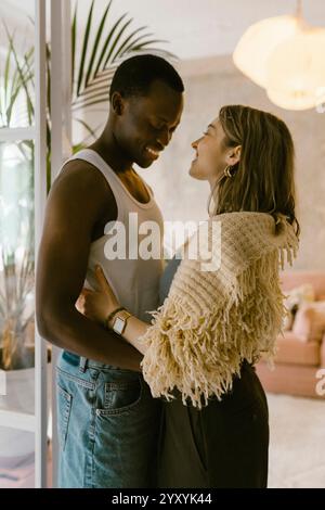 Seitenansicht eines lächelnden, multirassischen jungen Paares, das zuhause nebeneinander steht Stockfoto