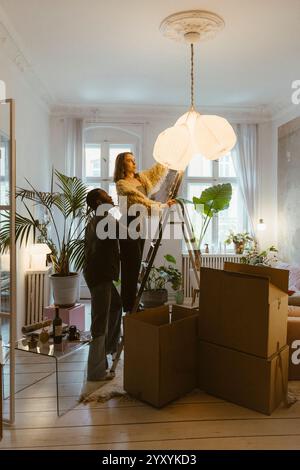 Junger Mann, der der Freundin hilft, auf einer Leiter zu stehen, während er die Pendelleuchte in der Wohnung aufhängt Stockfoto