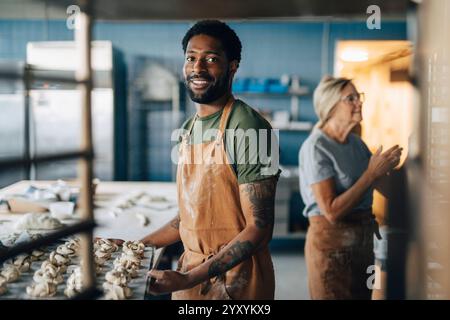 Porträt eines lächelnden männlichen Bäckers, der ein Tablett mit frischen Zimtbrötchen hält, während er in der Bäckerei steht Stockfoto