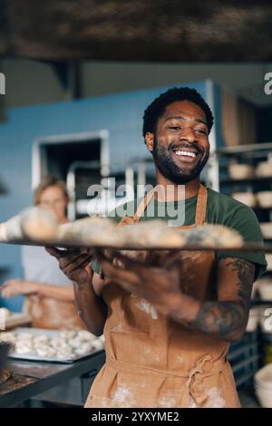 Glücklicher männlicher Bäcker, der ein Tablett mit frisch gebackenen Brötchen hält, während er in der Bäckerei arbeitet Stockfoto