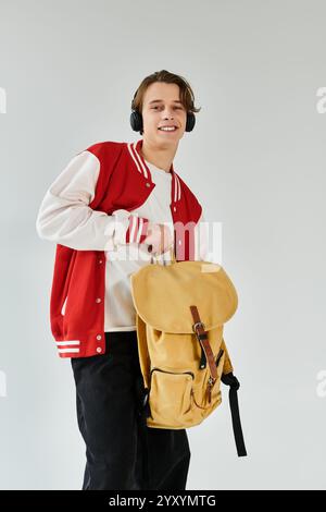 Ein hübscher Student mit einer trendigen Bomberjacke lächelt, während er im Studio einen Rucksack hält. Stockfoto