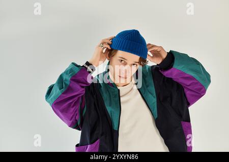 Ein gutaussehender junger Mann passt seine blaue Mütze an, während er im Studio eine lebhafte Bomberjacke trägt. Stockfoto