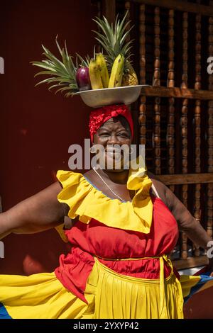 Lächelnde Kolumbianische Frau Mit Traditionellem Kleid Und Ausbalancierenden Tropischen Früchten Auf Dem Kopf Vor Der Roten Wand. Stockfoto