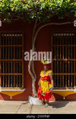 Eine Palenquera-Frau in Einem bunten Kleid mit Einem Obstkorb auf dem Kopf, die vor Einem Kolonialgebäude mit Holzfenstern steht Stockfoto