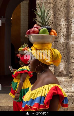 Ein Seitenprofil Einer Palenquera-Frau in Einem gelben und roten Kleid, die Einen Obstkorb auf dem Kopf in der Nähe Einer Steinmauer balanciert Stockfoto