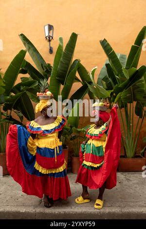 Zwei Palenquera-Frauen in hellen traditionellen Kleidern, die ihre Röcke halten und vor Palmen und Einer gelben Mauer lächeln. Stockfoto