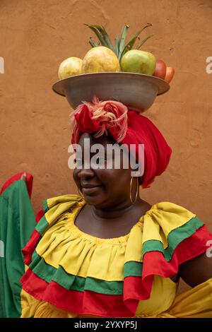 Eine Nahaufnahme Einer lächelnden Palenquera-Frau in Einem traditionellen roten und gelben Kleid, die Einen Obstkorb auf dem Kopf ausbalanciert. Stockfoto