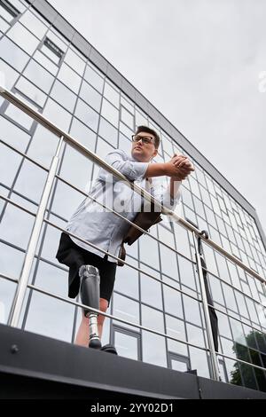 Ein junger Mann mit einer Beinprothese steht auf einem Balkon mit Blick auf die Stadt, verloren in Gedanken. Stockfoto