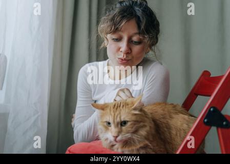 Junge Frau, die lustige Gesichter macht, während sie ihre Ingwerkatze streichelt, die zu Hause auf ihrem Schoß sitzt und Liebe und Zuneigung zu ihrem Haustier zeigt Stockfoto