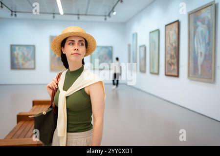 Mitten im Bild einer eleganten jungen kaukasischen Frau mit Strohhut, die in der Kunstgalerie posiert. Kopierbereich. Kulturerziehung und Museumstag. Stockfoto