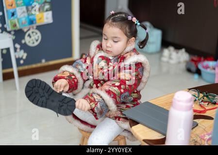 Ein kleines chinesisches Mädchen in traditioneller Tracht zieht Stiefel an. Sie ist bereit, das chinesische Neujahr zu feiern. Stockfoto