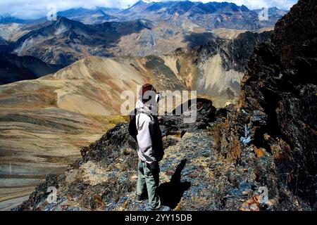 Ein Wanderer in den bolivianischen Anden steht auf einem Felsvorsprung in den bolivianischen Anden, der auf die fernen Berge zeigt. Verfügt über raues Gelände. Stockfoto