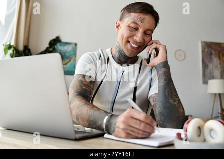 Ein hübscher junger Mann mit Tätowierungen macht einen Anruf, während er sich Notizen an seinem Schreibtisch notiert. Stockfoto