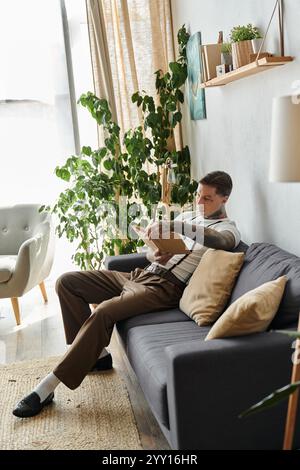 Hübscher junger Mann mit Tätowierungen sitzt bequem auf einer Couch und liest drinnen ein Buch. Stockfoto