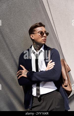 Hübscher junger Mann mit Tätowierungen steht mit überkreuzten Armen, elegant gekleidet in einer urbanen Umgebung. Stockfoto