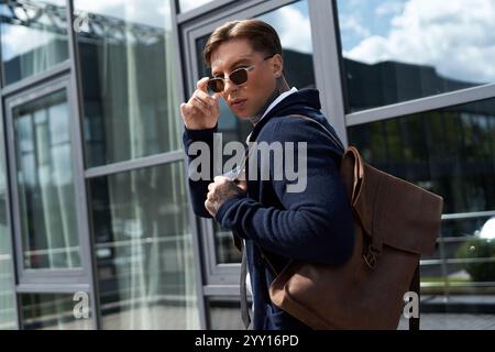 Ein junger Mann mit Sonnenbrille zeigt seine Tätowierungen, während er seine Tasche draußen verstellt. Stockfoto