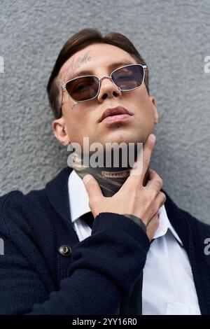 Mann mit Tattoos zeigt eine entspannte Pose vor einer grauen Wand, mit trendigen Sonnenbrillen und Kleidung. Stockfoto