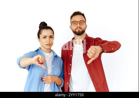 Unzufriedener arabischer Mann und kaukasische Frau mit Daumen nach unten, Ausdruck von Missbilligung und Enttäuschung, lässig gekleidet, stehen auf isoliertem weißen Hintergrund und schauen in die Kamera Stockfoto