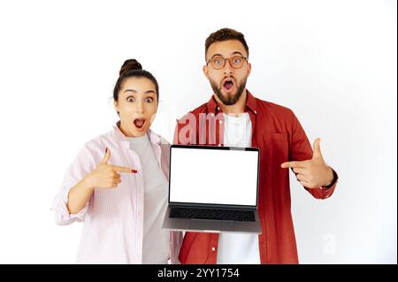 Überraschte Männer und Frauen arabischer oder kaukasischer Herkunft, die einen Laptop mit einem leeren Bildschirm halten, darauf zeigen und Staunen ausdrücken, Inhalte oder Produkte bewerben, stehen auf isoliertem weißen Hintergrund Stockfoto