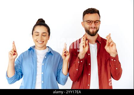 Hoffnungsvolle Mann und Frau, die mit geschlossenen Augen die Finger kreuzen und viel Glück wünschen, gekleidet in lässigen roten und blauen Hemden, stehen auf isoliertem weißem Hintergrund Stockfoto