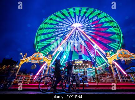 Schwerin, Deutschland. Dezember 2024. Besucher stehen vor einem Riesenrad auf dem Weihnachtsmarkt am Ufer des Pfaffenteichs. (Foto mit langer Belichtungszeit) in Mecklenburg-Vorpommern enden die meisten großen Weihnachtsmärkte wenige Tage vor Heiligabend - nur in Schwerin und Stralsund können die Glühweinstände und Mutzen-Stände noch nach den Feiertagen besucht werden. Der Schweriner Markt endet am 30. Dezember, der Stralsunder Markt endet erst im Dezember 31. Quelle: Jens Büttner/dpa/Alamy Live News Stockfoto