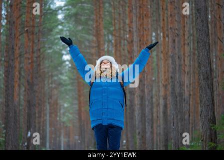 Wanderer mit blauer Daunenjacke und weißer Kapuze hebt sich in einem verschneiten Winterwald freudig die Arme und genießt die Freiheit und frische Luft der ruhigen Landschaft Stockfoto