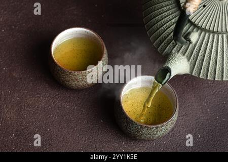 Frischer Grüntee in Keramikbecher gießen. Traditionelle japanische chinesische Teekanne aus Eisen und Teetassen auf dunkelbraunem Tisch aus nächster Nähe. Asiatische Teekultur. Stockfoto