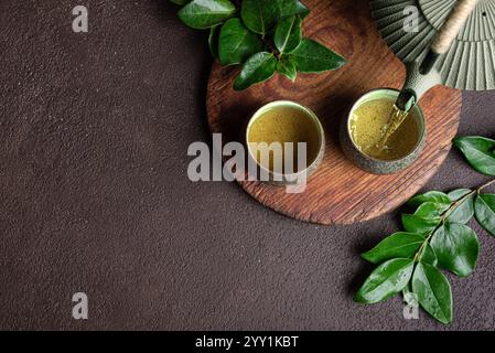 Asiatische Teekanne, die grünen Tee in Keramikbecher gießt. Traditioneller japanischer chinesischer Tee auf dunkelbraunem Hintergrund mit grünen Blättern, Draufsicht, Kopie Spac Stockfoto