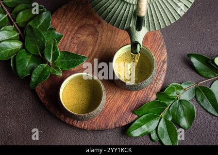 Asiatische eiserne Teekanne, die grünen Tee in Tassen gießt. Traditioneller japanischer chinesischer Tee auf dunkelbraunem Hintergrund mit grünen Blättern, Blick von oben. Aes Stockfoto
