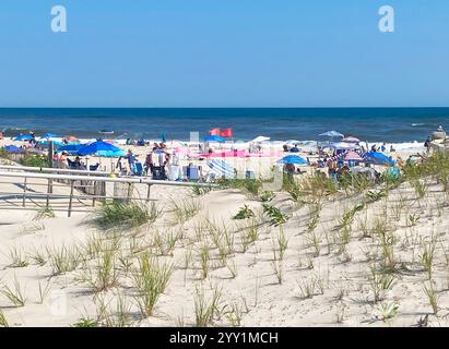 Babylon, New York, USA - 10. August 2024: Gruppen von Menschen genießen einen sonnigen Strandtag unter Sonnenschirmen mit Fahnen, die in der sanften Meeresbrise fliegen. Stockfoto