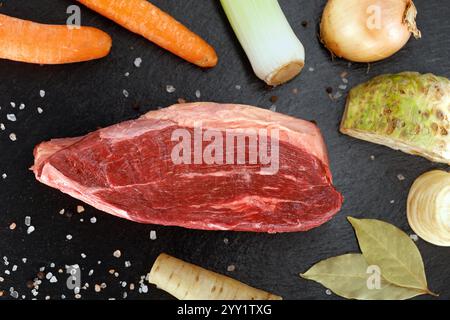 Frisches Stück rohes Rindfleisch aus der Kappe mit Wurzelgemüse auf schwarzem Schieferteller, Zutaten für die Zubereitung von gekochtem Rindfleisch, Draufsicht Stockfoto