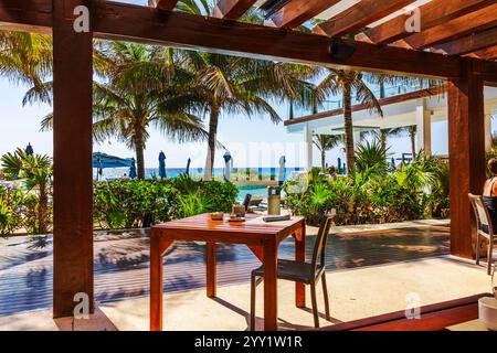 Außenterrasse unter hölzerner Pergola mit Tisch und Stühlen mit Blick auf den tropischen Garten, Palmen und das türkisfarbene Karibische Meer. Cancun. Mexiko. Stockfoto