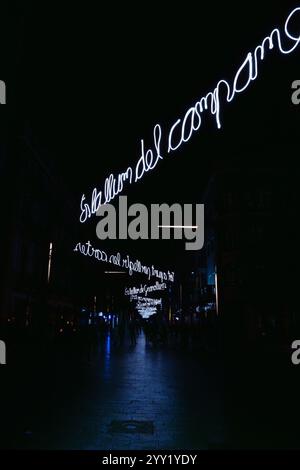 Dunkel beleuchtete Nachtstraße mit Texten im Zickzack-Neon-Hintergrund, die entlang der gesamten kopfsteingepflasterten Straße laufen, mit Silhouetten von Menschen, die entlang schlendern Stockfoto